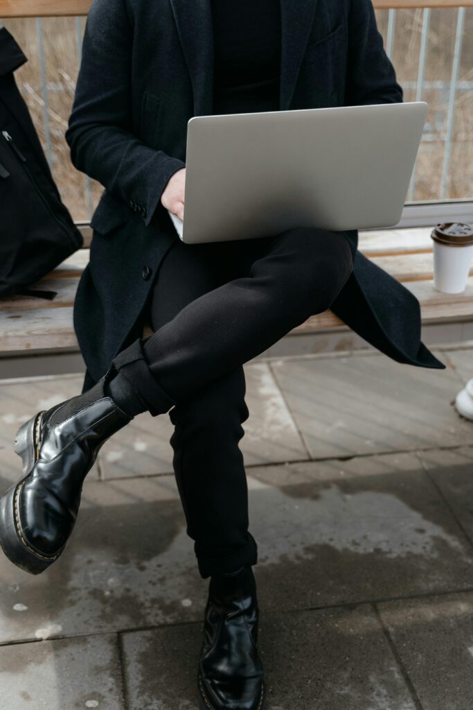 pexels-photo-7252538-7252538 Person in black coat sitting with a laptop on a wooden bench outdoors.